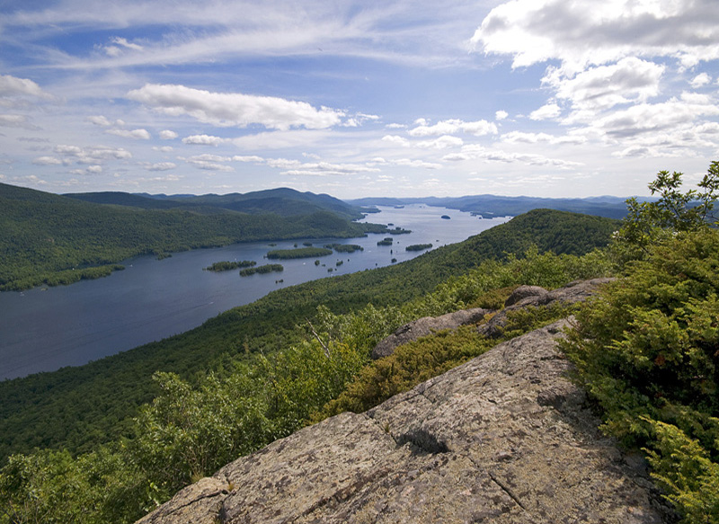 Scenic view of a large lake winding through lush green hills under a partially cloudy sky. The foreground features rocky terrain with scattered vegetation, and the distant horizon is filled with rolling mountains.