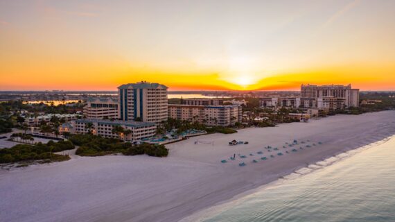 Sunset over a beach with sandy shores and calm ocean waves. A row of turquoise lounge chairs lines the beach. Several buildings and palm trees are visible, with a vibrant orange and yellow sky in the background.