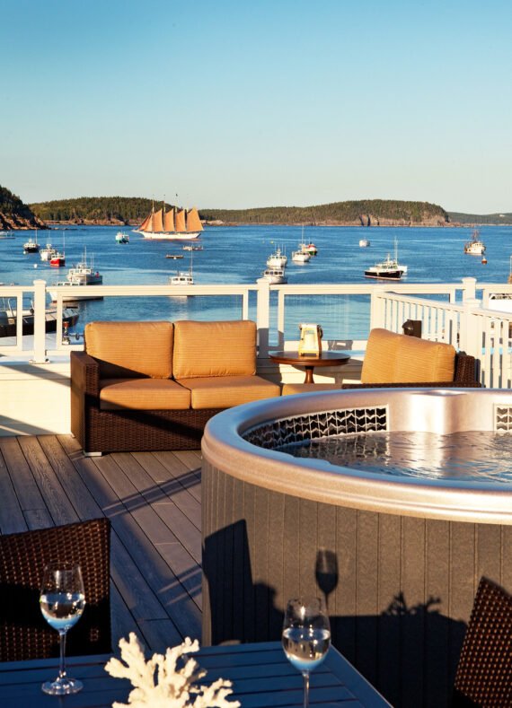 A rooftop terrace with a hot tub and seating area overlooks a scenic harbor. Boats are on the water, with a large sailboat in the background. Two glasses of white wine are on a table, and the view extends to distant islands under a clear sky.