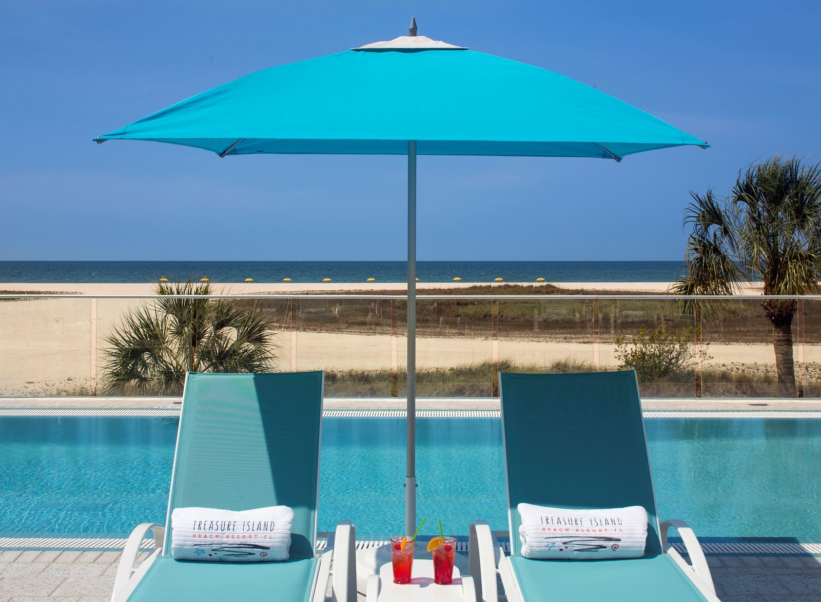 Two lounge chairs with Treasure Island towels sit under a blue umbrella beside a pool. Two colorful drinks are on a table between the chairs. The beach and ocean are visible in the background under a clear blue sky.