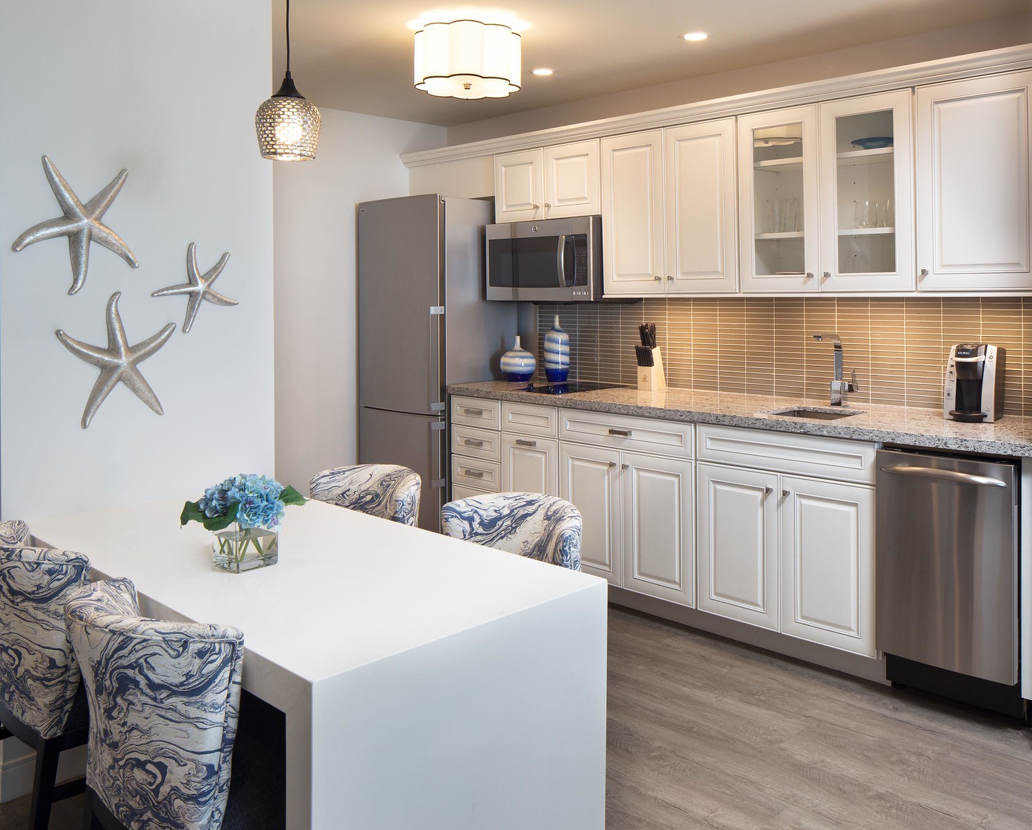 Modern kitchen with white cabinets and a granite countertop. A stainless steel fridge and dishwasher are installed. A white dining table with blue-patterned chairs is in the foreground. Starfish decor adorns the wall, and a pendant light hangs above.