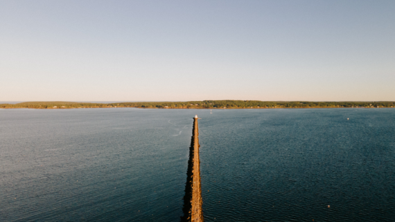 Aerial view of a long, straight breakwater extending into calm, blue water. A small lighthouse stands at the end of the breakwater. The horizon shows a distant shoreline with trees under a clear sky.