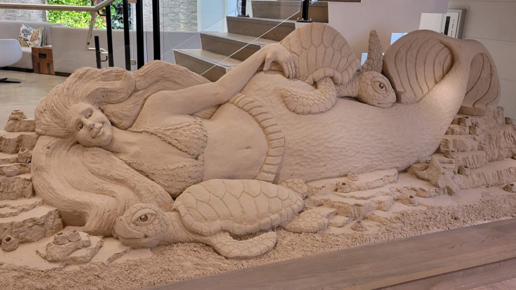 A detailed sand sculpture depicting a mermaid surrounded by sea turtles. The mermaid is reclining with flowing hair, and the turtles appear to interact with her. The sculpture is set indoors near stairs.