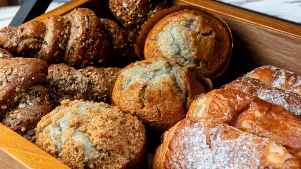 A wooden tray filled with various pastries: seed-encrusted rolls, blueberry muffins, crumb-topped muffins, and flaky pastries dusted with powdered sugar, creating an inviting and delicious assortment.