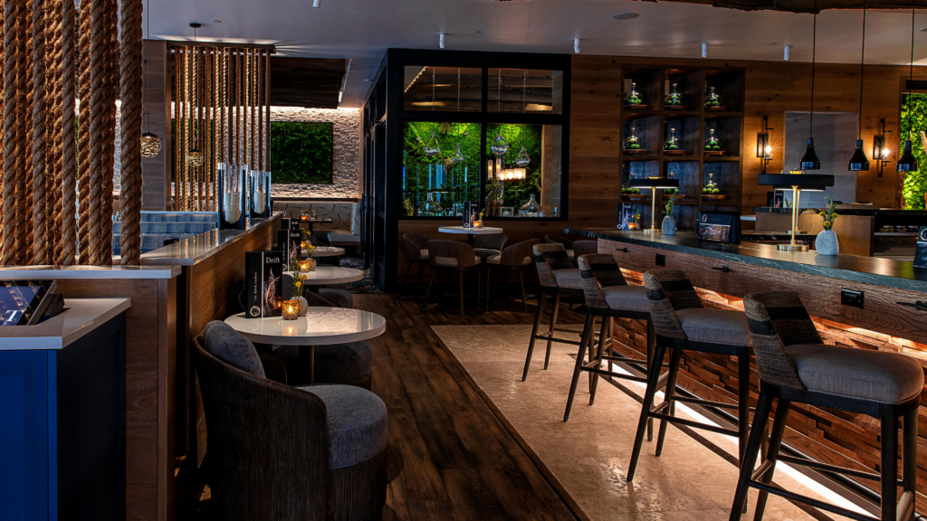 A modern, dimly lit bar with wooden decor. High-backed bar stools line the counter, and small tables with cushioned chairs are set in the seating area. Hanging pendant lights add warmth, and greenery is visible through large windows.
