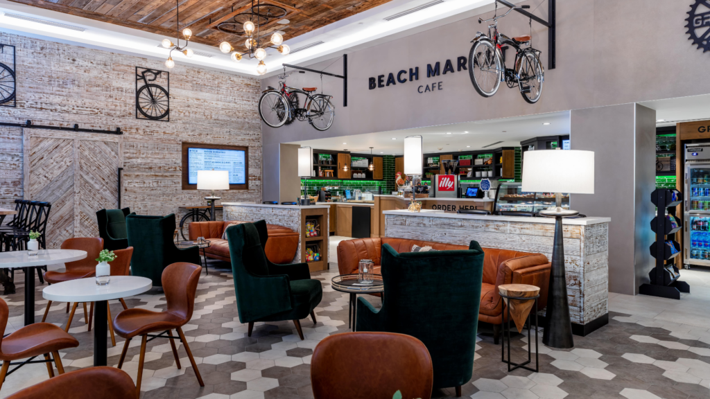 Coffee shop interior with a rustic theme. Features wooden accents, bicycles hung on the wall, and a combination of green and brown seating. Hexagonal floor tiles and a large overhead light fixture add to the ambiance. A sign reads Beach Market Cafe.