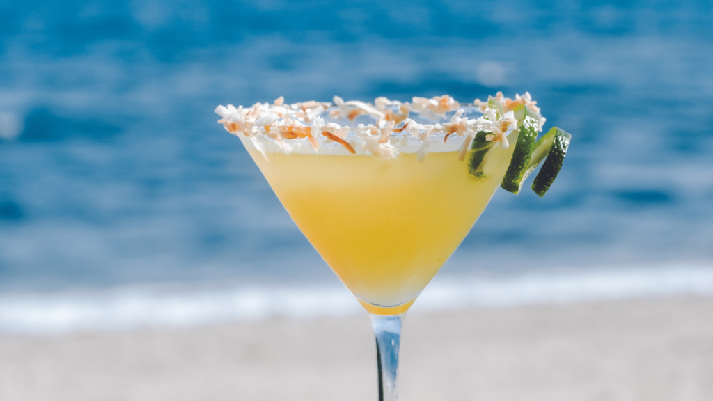 A tropical cocktail in a martini glass, garnished with lime slices and a coconut rim, against a blurred ocean background.