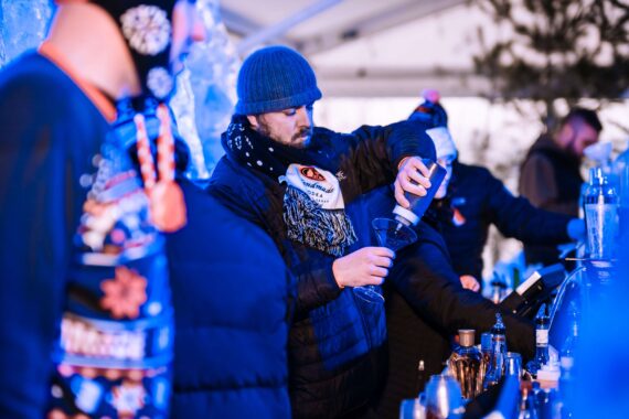 A bartender wearing winter clothing pours a drink into a glass at an outdoor bar. The scene has a cold, festive atmosphere with blue lighting and other people in warm attire in the background.