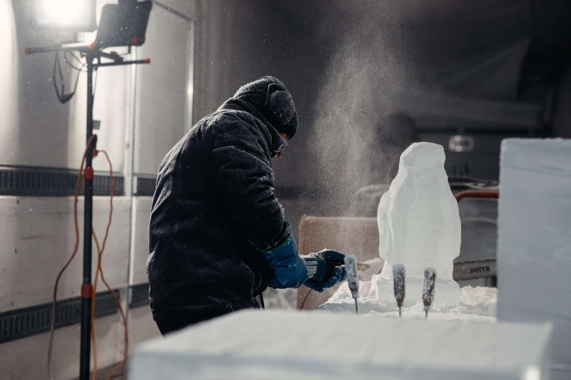 A person in winter clothing sculpts an ice block using a tool, with shavings flying around. They are indoors, working on a detailed sculpture, with a light source illuminating the scene.