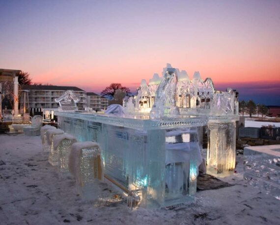 A stunning ice sculpture of a bar with ornate details, set against a colorful sunset sky. The frozen structure glows with subtle lighting, creating a magical atmosphere. Snow covers parts of the ground around the sculpture.