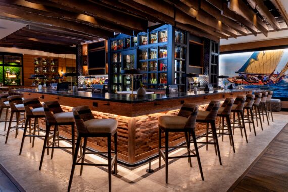 A modern, spacious bar with a wooden interior features a large, illuminated central bar counter surrounded by tall bar stools. Behind the bar, shelves display a variety of bottled drinks. A nautical-themed painting adorns the wall.