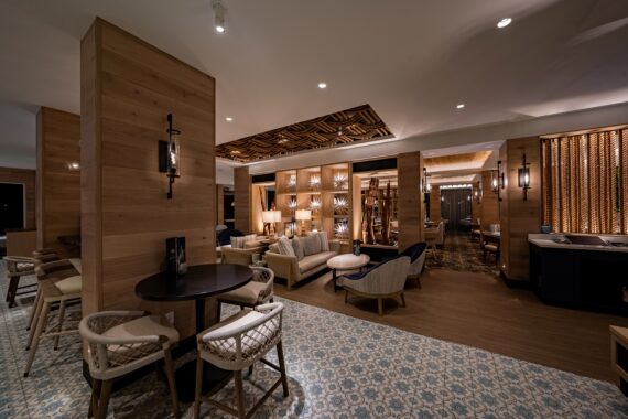 A cozy, dimly lit lounge area with modern wooden decor, featuring comfortable chairs and sofas, patterned flooring, and a decorative ceiling. Bookshelves and soft lighting create a warm, inviting atmosphere.