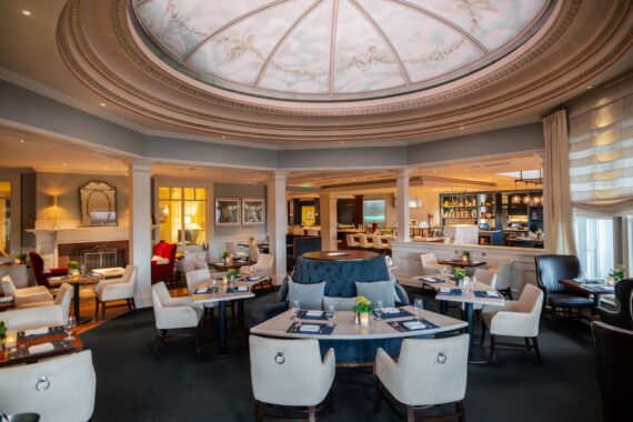 A spacious, elegant restaurant with a domed ceiling painted with clouds and sky. The room features a central circular seating area, surrounding tables, plush chairs, and warm lighting. Tables are set with placemats and vases of fresh flowers.