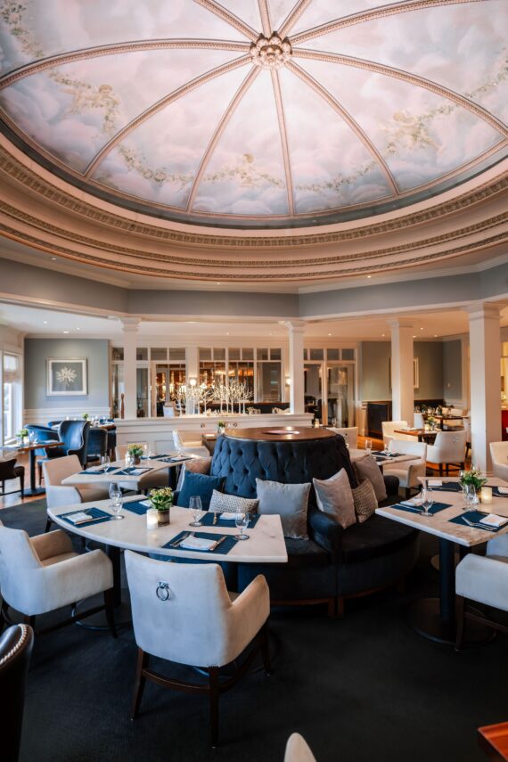 A luxurious restaurant with a grand, painted dome ceiling, white walls, and floor-to-ceiling windows. The elegant seating arrangement features plush chairs and sofas around marble tables, adorned with flower vases. A stylish bar area is visible in the background.