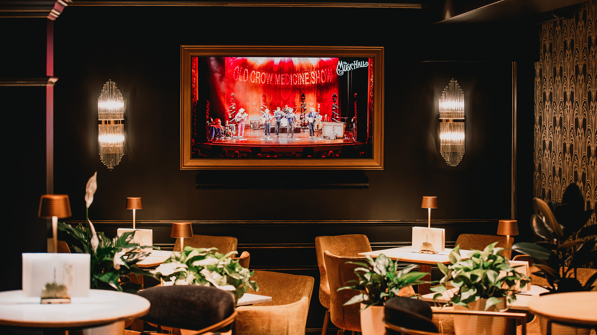 A cozy, dimly lit lounge with brown chairs, round tables, potted plants, and wall-mounted lights. A large screen on the wall displays musicians performing on a red-lit stage.