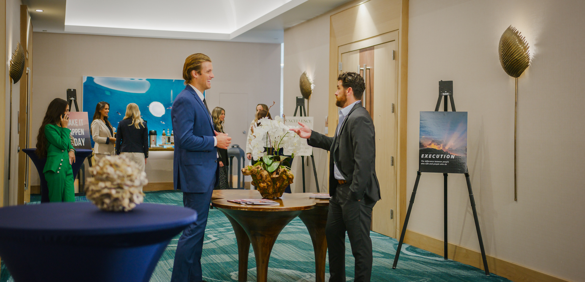 Two men in suits converse near a table with a plant in a professional setting. Other attendees are mingling in the background. Posters and decorative elements are visible on the walls.