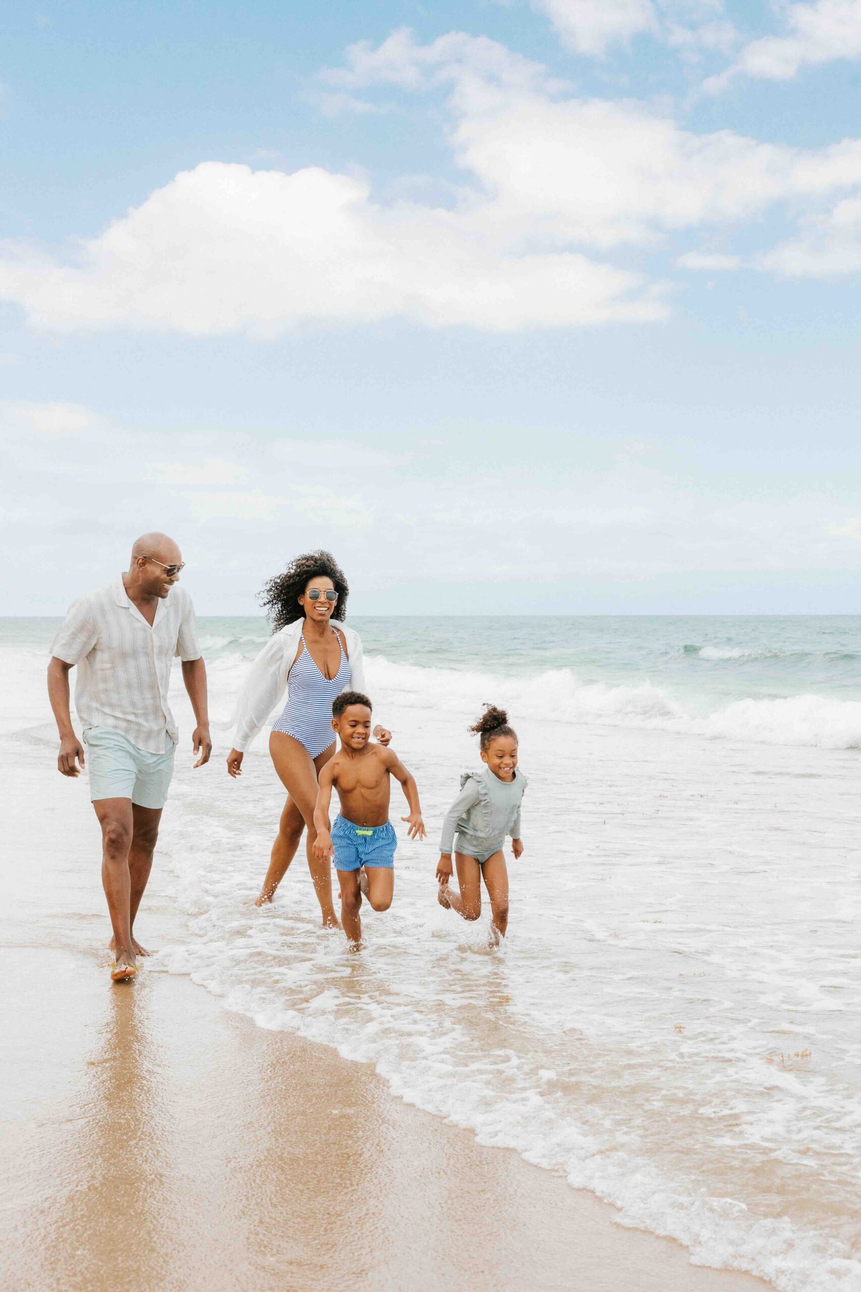 A family of four enjoys a day at the beach. Two adults and two children walk along the shore, with the ocean waves gently lapping at their feet. The sky is bright with scattered clouds. Everyone is dressed in light, beach-appropriate clothing.