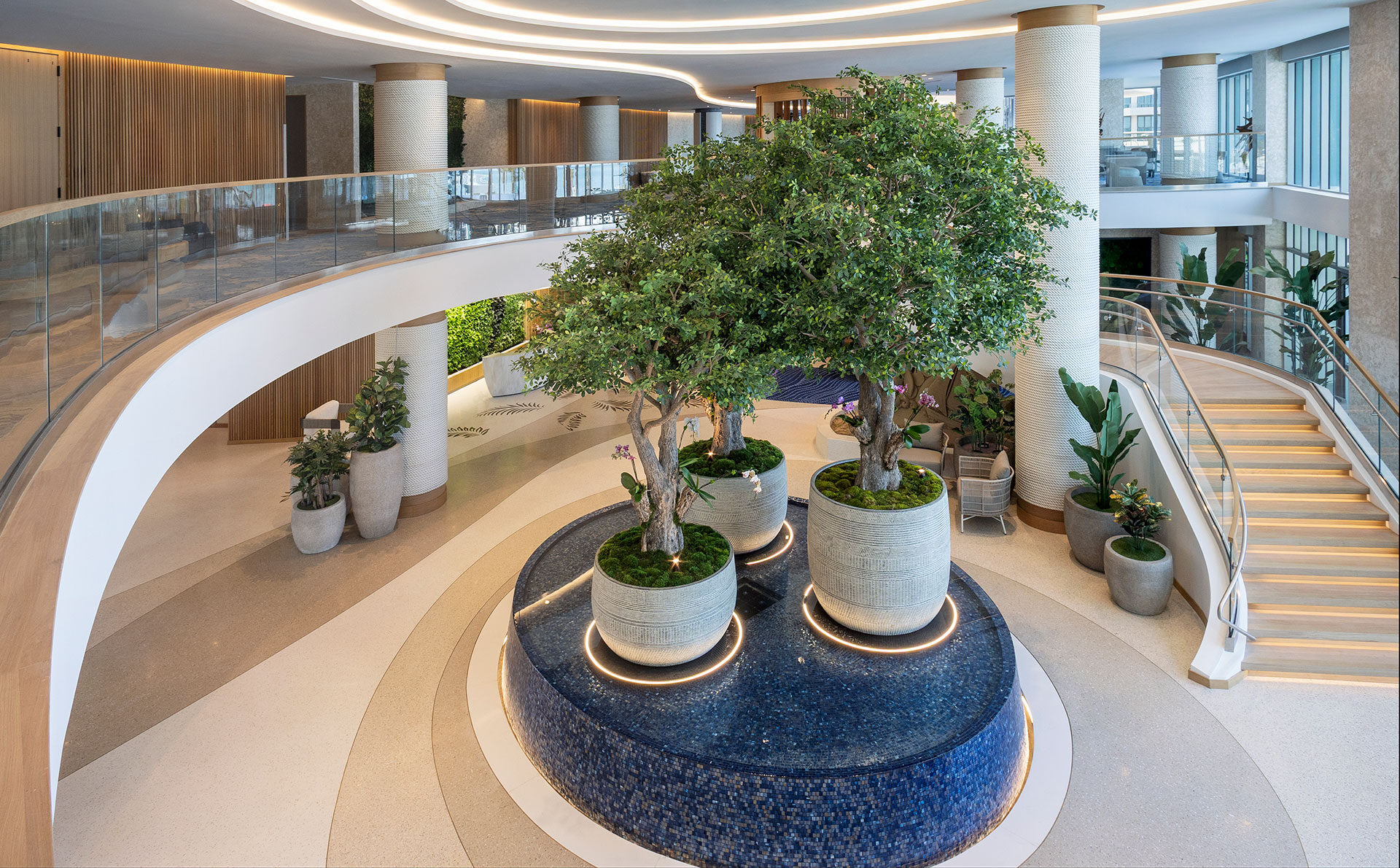 A modern, spacious indoor atrium at new hotels in Clearwater Beach, Florida, featuring large potted trees on a circular blue-tiled platform, curved staircases, glass railings, and abundant natural light from expansive windows.