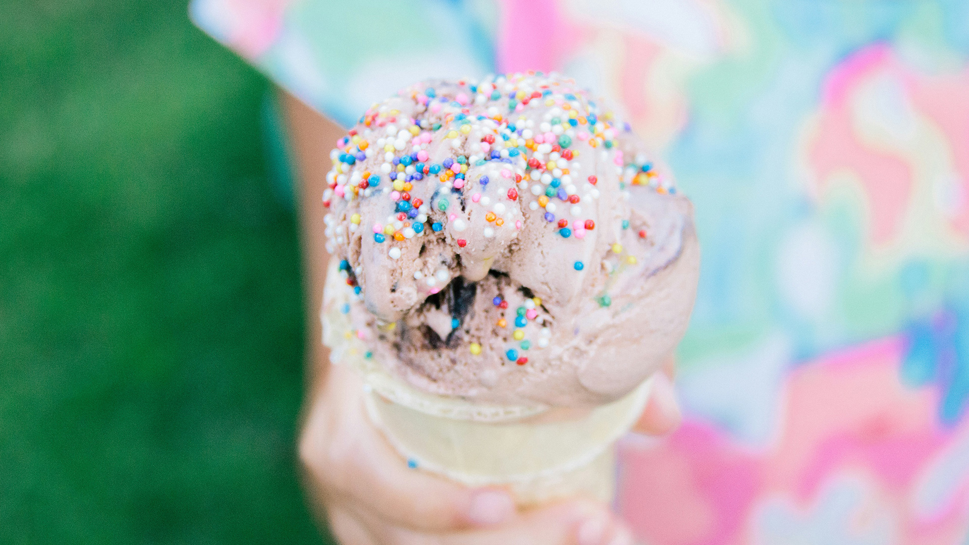 A hand holds a chocolate ice cream cone topped with colorful sprinkles. The background is blurred, showing part of a person wearing a pastel, patterned shirt and green grass.