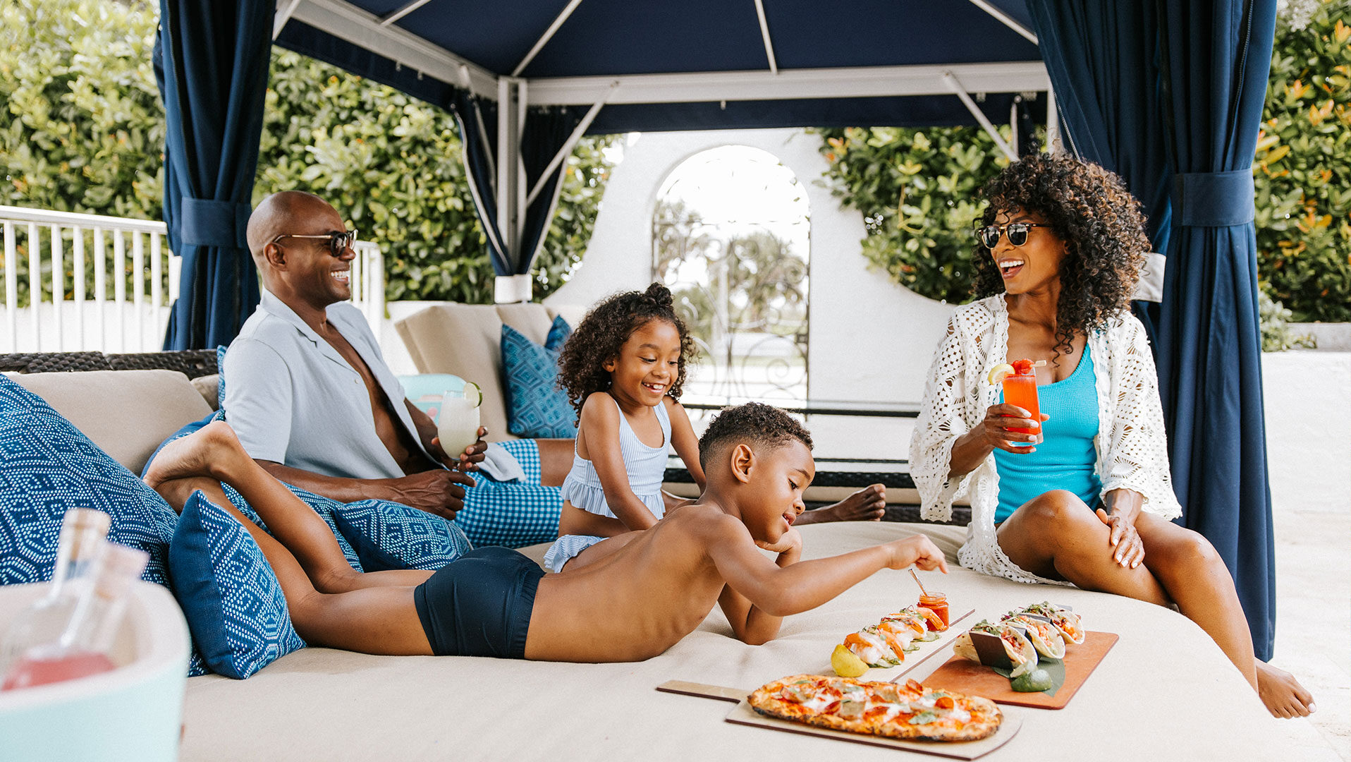 A family of four relaxes in a shaded outdoor cabana, smiling and enjoying drinks and snacks. Two children in swimwear reach for food on a table while the parents sit nearby, chatting and laughing.