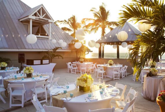 An elegant outdoor wedding reception with round tables adorned with white chairs and floral centerpieces. String lights and hanging lanterns create a festive atmosphere. Palm trees and a sunset sky enhance the tropical setting.