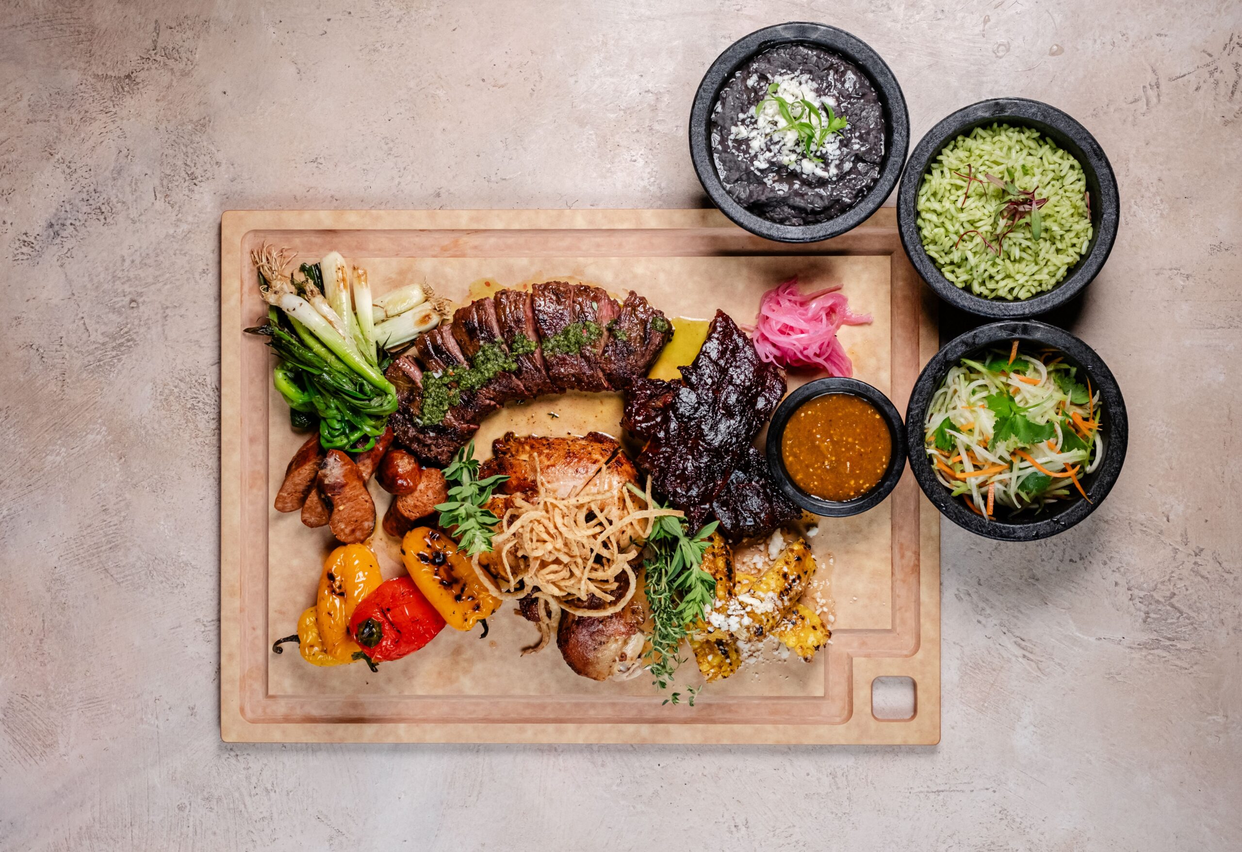 A wooden board with a colorful assortment of grilled meats, vegetables, and garnishes, including peppers, onions, and herbs. Accompanied by three bowls containing black beans, green rice, and a salad with dressing.
