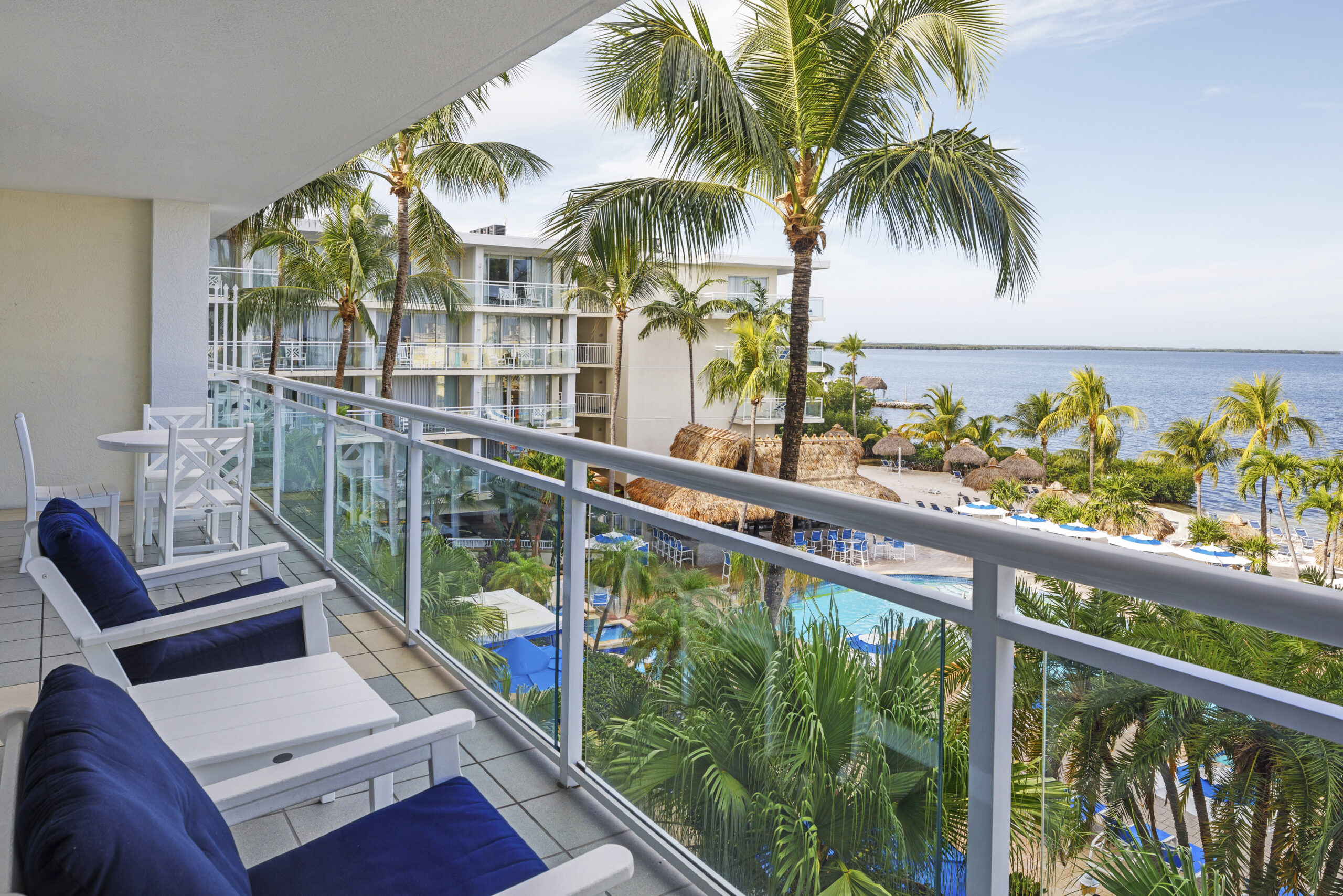 A hotel balcony with two chairs and a table overlooks a tropical resort. Palm trees, a pool, and thatched-roof structures are visible below. The ocean stretches out under a clear blue sky in the background.