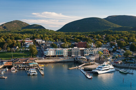 Aerial view of a coastal town with a marina, boats docked on clear blue water, a cluster of buildings and houses, and green mountains in the background under a clear sky.