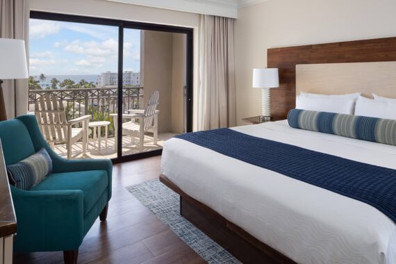 Hotel room with a king-sized bed adorned with white and blue linens, near a turquoise armchair. A large window leads to a balcony with chairs, offering a view of palm trees and a distant cityscape under a blue sky.