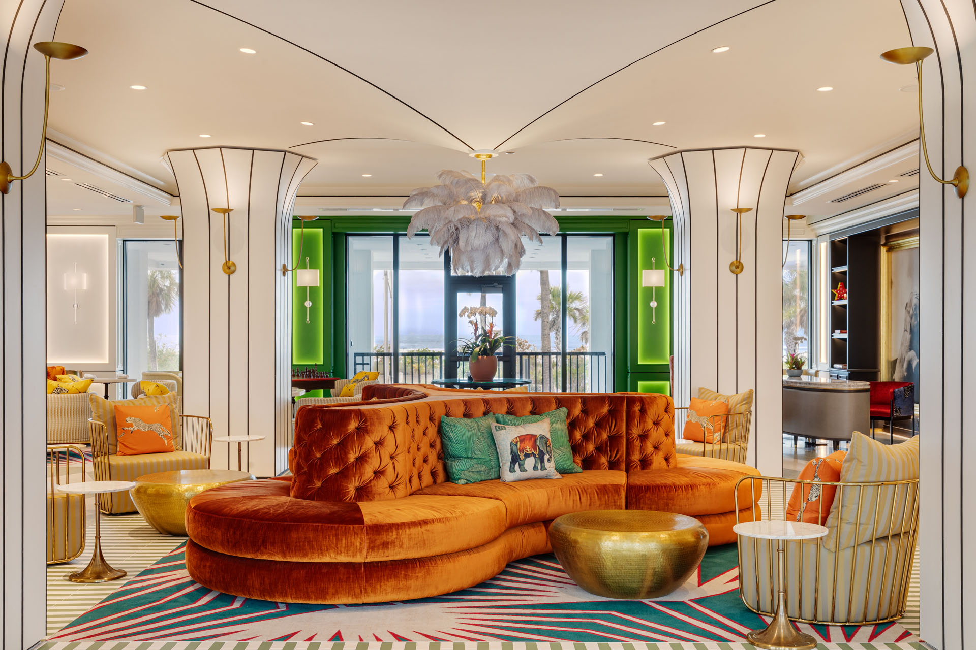 A stylish hotel lounge with a curved orange velvet sofa, colorful cushions, gold round tables, palm-themed decor, white feather chandelier, and green accent walls, with large windows offering a view of palm trees outside.