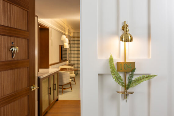 Hotel room entrance featuring a door slightly open, revealing a stylish interior with seating. A golden sconce with room number 104 and decorative fern adorns the wall. Warm lighting creates an inviting atmosphere.