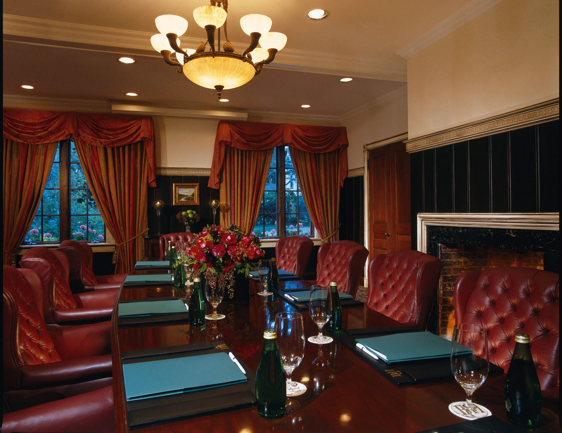 A luxurious conference room with a polished wooden table surrounded by red leather chairs. The table is set with green folders, water bottles, and glasses. Elegant drapes and floral arrangements add a sophisticated touch. A chandelier lights the room.