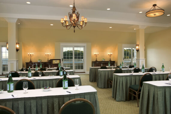 A well-lit conference room features rows of tables covered with white and green tablecloths, each set with water bottles, glasses, and notepads. Elegant chandeliers and wall sconces illuminate the space, which includes large windows overlooking greenery.