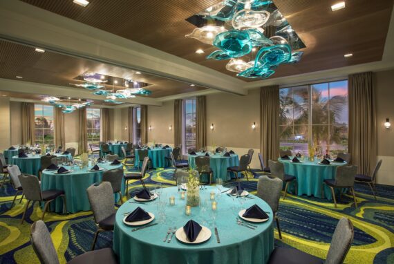 An elegantly set banquet hall with round tables covered in teal tablecloths and blue napkins. Modern glass light fixtures hang from the ceiling. Large windows offer a view of palm trees, with patterned carpet adding a vibrant touch.