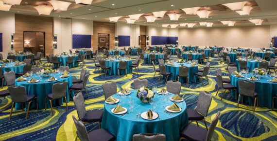 A banquet hall set up for an event. Round tables covered in turquoise tablecloths are arranged neatly, each with folded napkins, cutlery, and floral centerpieces. The floor features a blue and green patterned carpet, and the ceiling has decorative lights.