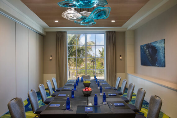 A modern conference room with a long rectangular table, surrounded by twelve gray chairs. Blue glass pendant lights hang above. The table holds blue water bottles, notepads, and a bowl of fruit. A large window reveals tropical palm trees outside.